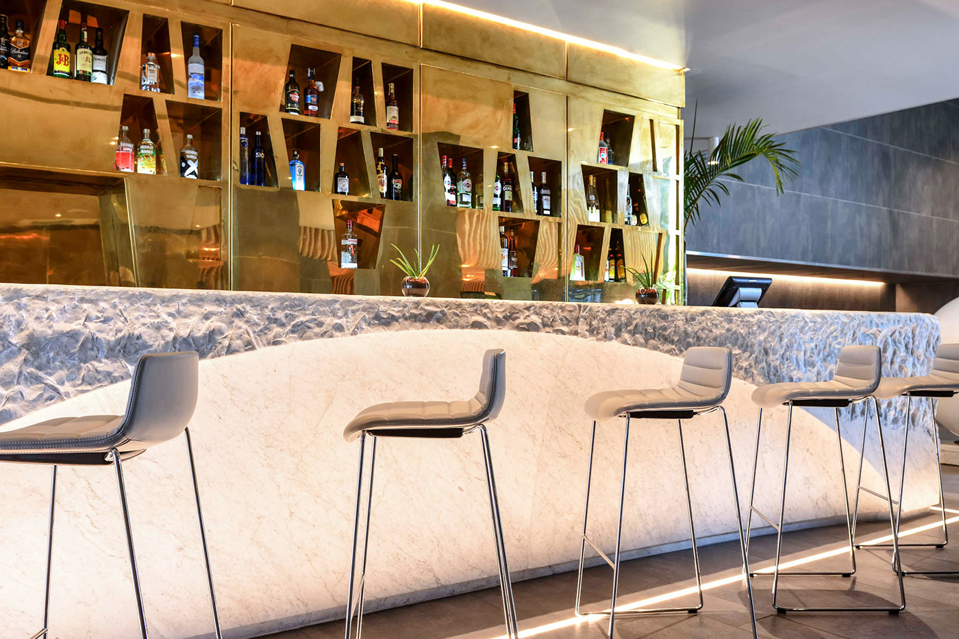 Un bar-boutique moderne avec une élégante étagère aux lignes dorées remplie de diverses bouteilles d'alcool. La façade du bar est recouverte de marbre blanc avec un motif texturé. Quatre grands tabourets de bar blancs avec des sièges rembourrés sont soigneusement alignés contre le comptoir. Une plante est visible en arrière-plan.