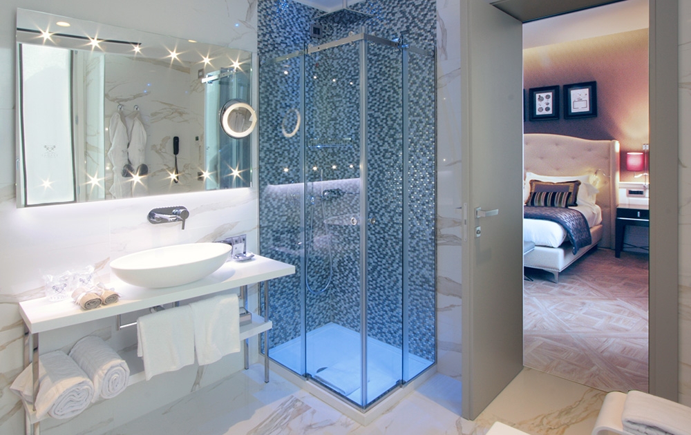 Salle de bains luxueuse aux murs et au sol en marbre, dotée d'une cabine de douche en verre moderne avec carrelage bleu et d'un grand miroir avec éclairage de vanité. Un lavabo blanc sur un plan de travail et des serviettes soigneusement pliées ajoutent à l'élégance. La porte s'ouvre sur une chambre bien éclairée avec une literie moelleuse, des œuvres d'art et des touches d'ameublement hôtelier.