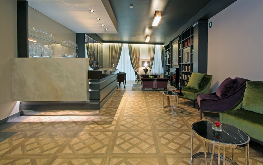 Le hall d'un hôtel-boutique moderne est doté d'un parquet à motifs, d'un bureau de réception à gauche et d'un coin salon avec des chaises en velours vert et violet à droite. L'espace respire le luxe avec des plafonniers bien éclairés, de grandes fenêtres ornées de rideaux transparents et des étagères chics en arrière-plan.