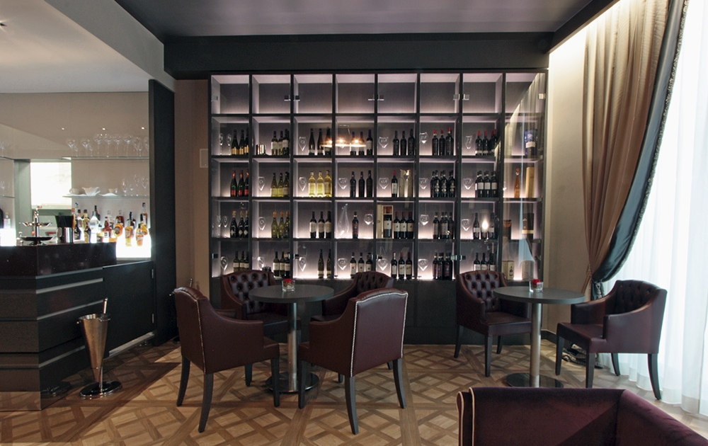 Un espace salon moderne avec un bar élégant dégageant une ambiance luxueuse. Quatre tables rondes sont entourées de chaises en cuir marron. Derrière les tables, il y a une grande étagère avec rétroéclairage mettant en valeur diverses bouteilles. Le sol présente un motif géométrique et des rideaux bordent le côté droit.