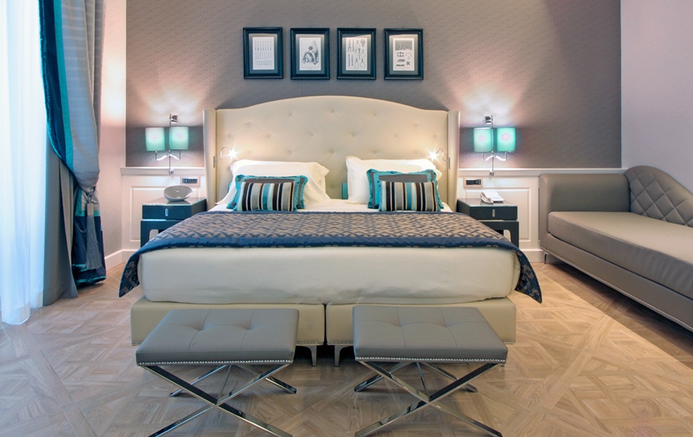 Une chambre d'hôtel moderne dotée d'un grand lit bien fait avec une tête de lit capitonnée, flanquée de deux tables de nuit et de petites lampes vertes. Au pied du lit se trouvent deux poufs. À droite se trouve une chaise longue. La chambre présente un agencement luxueux avec une palette de couleurs neutres et des œuvres d'art encadrées au-dessus du lit.