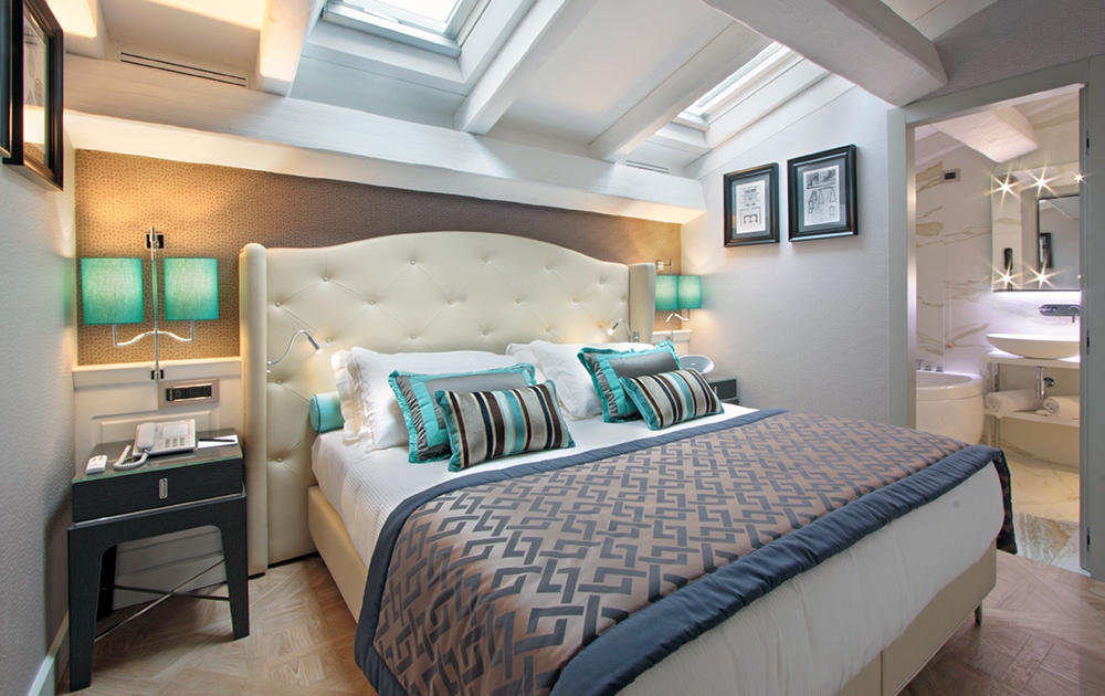 Une chambre confortable avec un lit double orné d'oreillers bleu sarcelle et blancs et d'une couette à motifs. La chambre comprend deux lampes de chevet bleu sarcelle, des œuvres d'art encadrées sur les murs et une lucarne. À droite, une porte s'ouvre sur une salle de bains moderne avec des miroirs éclairés.