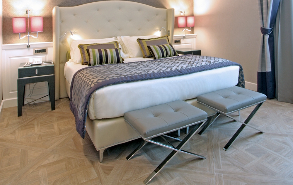 Chambre d'hôtel moderne dotée d'un grand lit à tête de lit capitonnée avec des oreillers à motifs et une couette texturée. Deux bancs élégants sur mesure avec des pieds métalliques se trouvent au pied du lit. Il y a deux tables de nuit avec des lampes de table et de grandes fenêtres avec des rideaux élégants sur le côté, complétant l'ambiance de boutique.