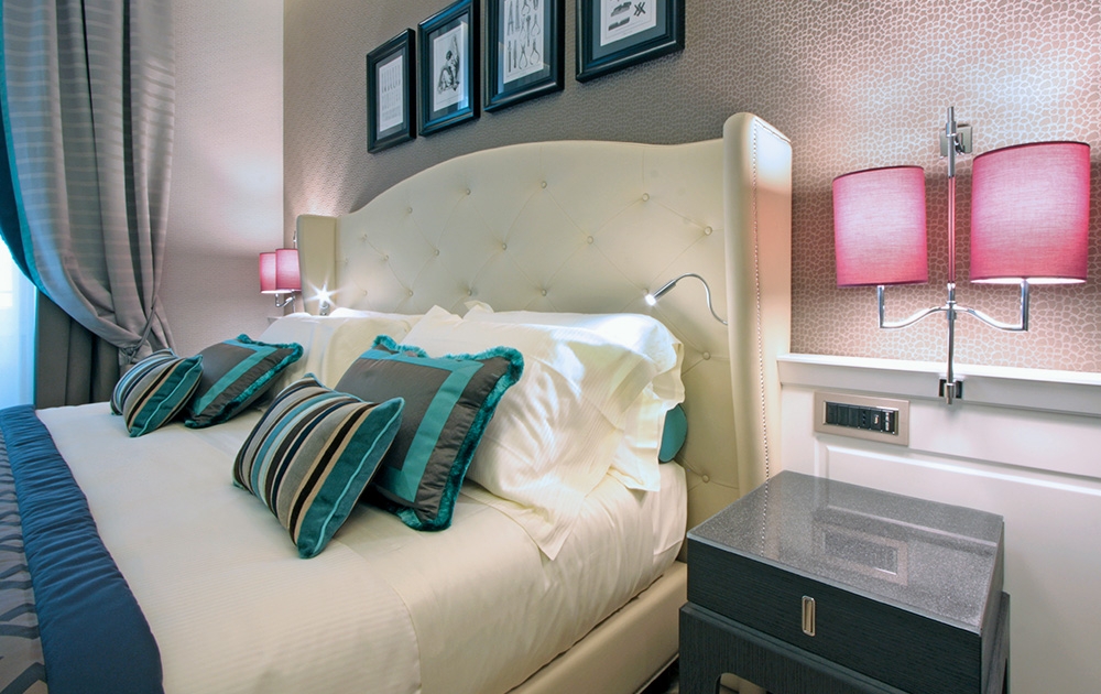 Une chambre moderne et bien éclairée avec une tête de lit moelleuse et capitonnée et un lit soigneusement fait orné d'oreillers bleu sarcelle et gris. Des tables de chevet avec des lampes roses sont disposées des deux côtés. Des œuvres d'art encadrées décorent le mur à la texture beige derrière le lit, mettant en valeur un agencement impeccable qui ressemble à un aménagement hôtelier.