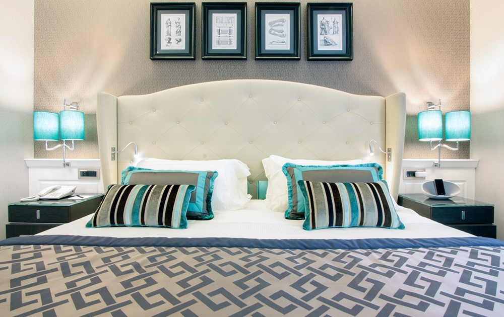 Une chambre d'hôtel-boutique moderne dispose d'une grande tête de lit capitonnée surmontée d'œuvres d'art encadrées. Le lit est orné d'oreillers et d'un couvre-lit à motifs géométriques. Les tables de nuit contiennent des lampes turquoise assorties aux oreillers décoratifs, et il y a un téléphone sur la table de nuit de gauche.