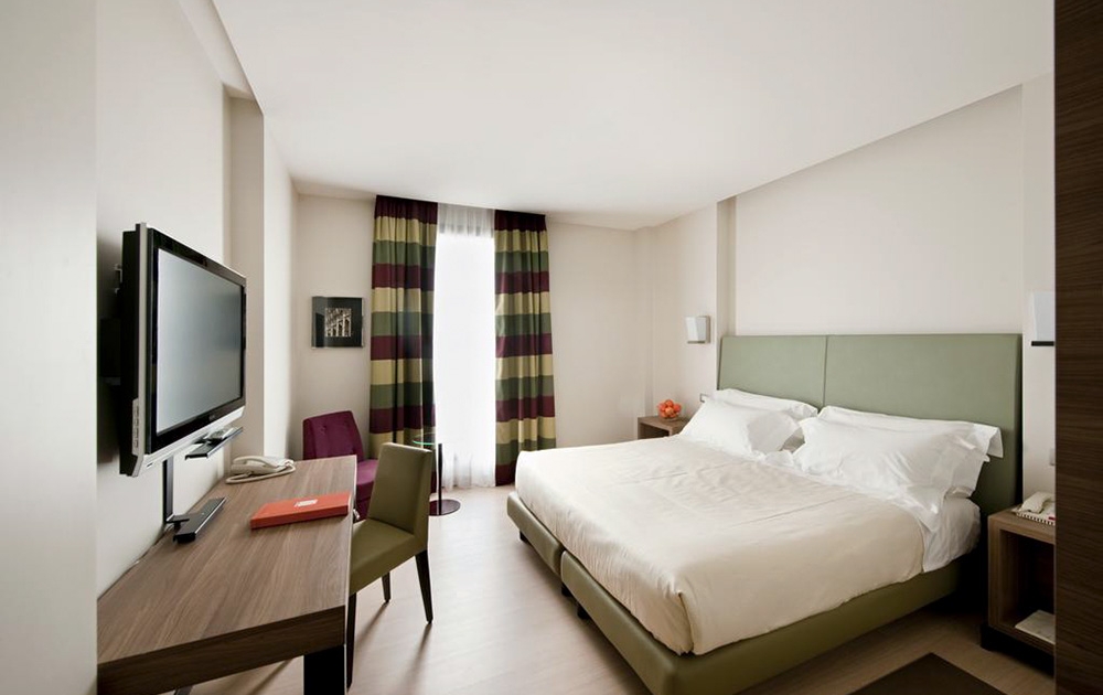 Chambre d'hôtel-boutique moderne avec un lit double avec une tête de lit tapissée de vert, une literie blanche et des tables de chevet. L'agencement de la chambre comprend une télévision murale, un bureau avec une chaise, une petite table ronde avec une chaise violette et une fenêtre avec des rideaux rayés.