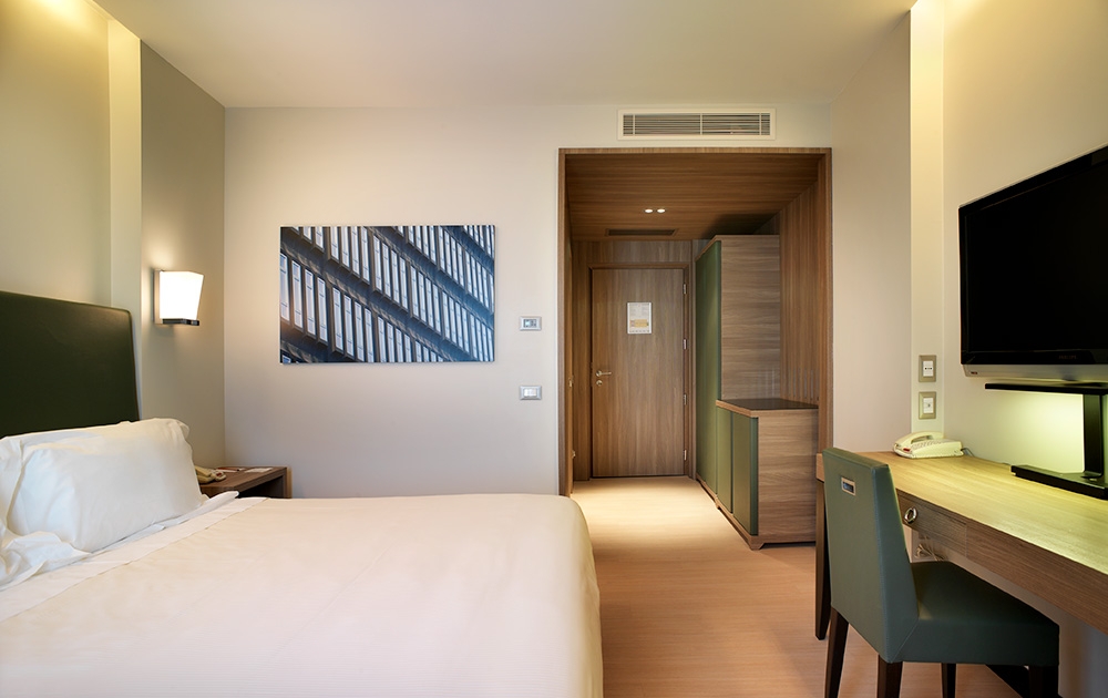 Une chambre d'hôtel moderne de style boutique avec un lit bien fait à gauche, avec une literie blanche et une tête de lit verte. Il y a une télévision à écran plat fixée au mur à droite, un bureau et une chaise, et une porte donnant sur le couloir d'entrée. Une œuvre d'art abstraite est accrochée au mur à côté du lit, mettant en valeur un agencement impeccable.