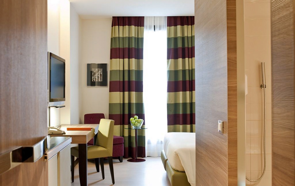 Une chambre d'hôtel moderne avec un agencement exquis, comprenant un lit, un petit bureau et une télévision à écran plat fixée au mur. La chambre offre des touches de luxe comme une grande fenêtre avec des rideaux multicolores à rayures vertes, rouges et beiges. Une chaise confortable et un petit bol de pommes vertes complètent le bureau.