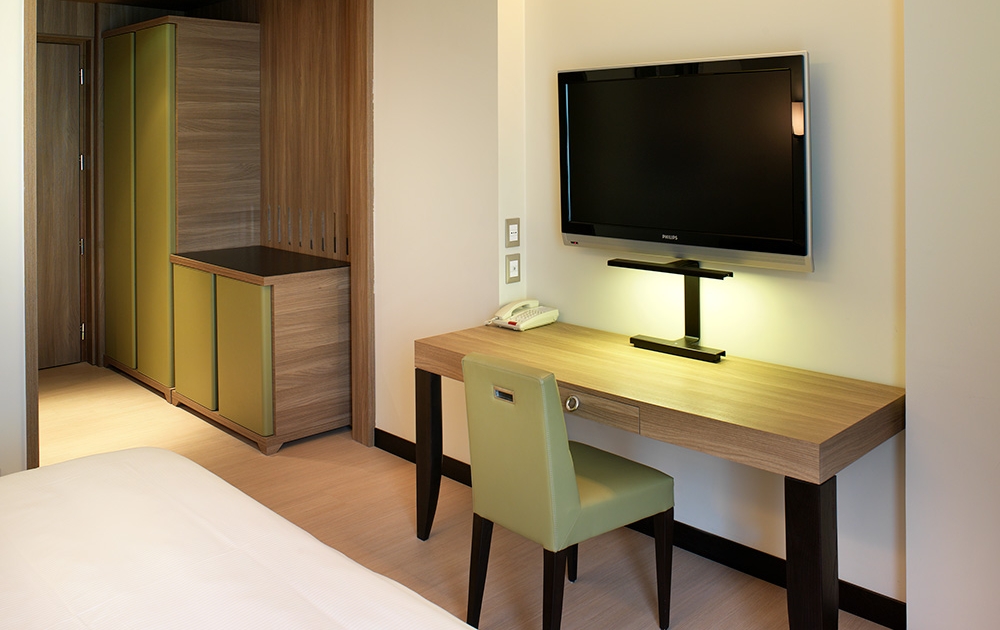 Une chambre d'hôtel dispose d'un téléviseur à écran plat fixé au mur au-dessus d'un bureau en bois avec une chaise verte, mettant en valeur l'aménagement hôtelier précis. Un téléphone et une lampe sont posés sur le bureau. À gauche se trouve une armoire fermée avec des portes vertes, complétant le design sur mesure. Le coin du lit est partiellement visible sur le côté gauche.