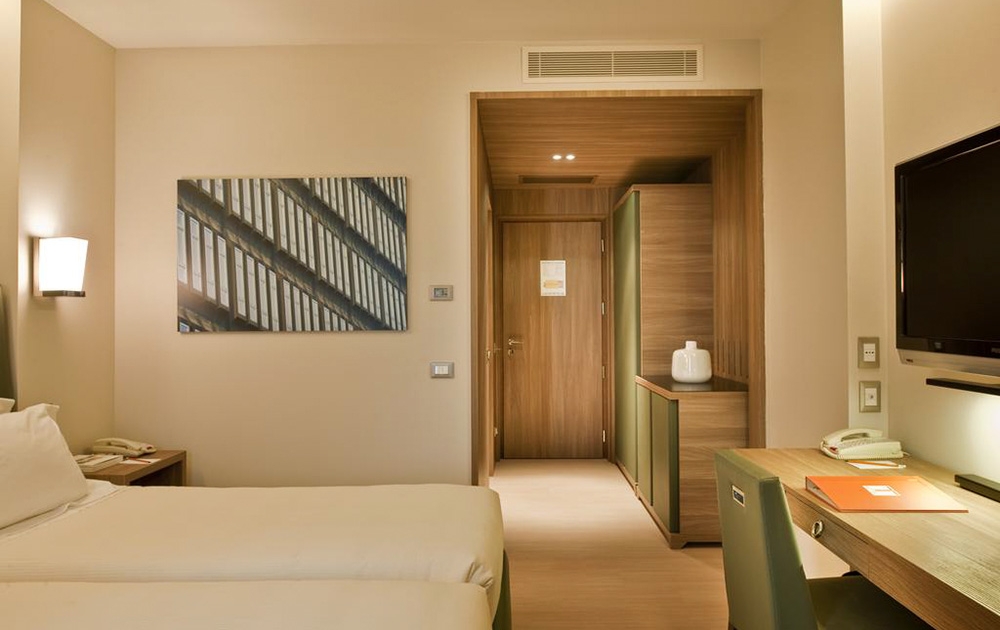 Une chambre d'hôtel-boutique moderne avec deux lits jumeaux soigneusement agencés contre un mur beige. Sur le mur au-dessus des lits se trouve une œuvre d'art abstraite encadrée. La chambre comprend un bureau en bois avec une chaise, une télévision murale et un meuble. La porte d'entrée est visible au fond, soulignant son agencement bien pensé.