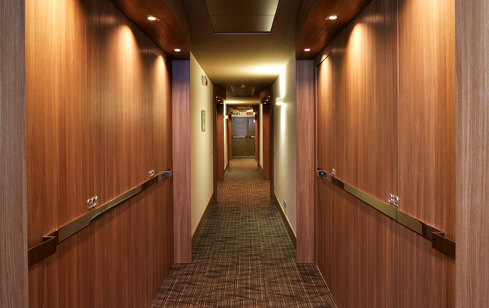 Un long couloir bien éclairé avec des murs lambrissés de bois et une moquette à motifs présente un agencement d'éclairage encastré au plafond et des rampes des deux côtés. Au fond, il y a une porte fermée flanquée d'élégants luminaires.
