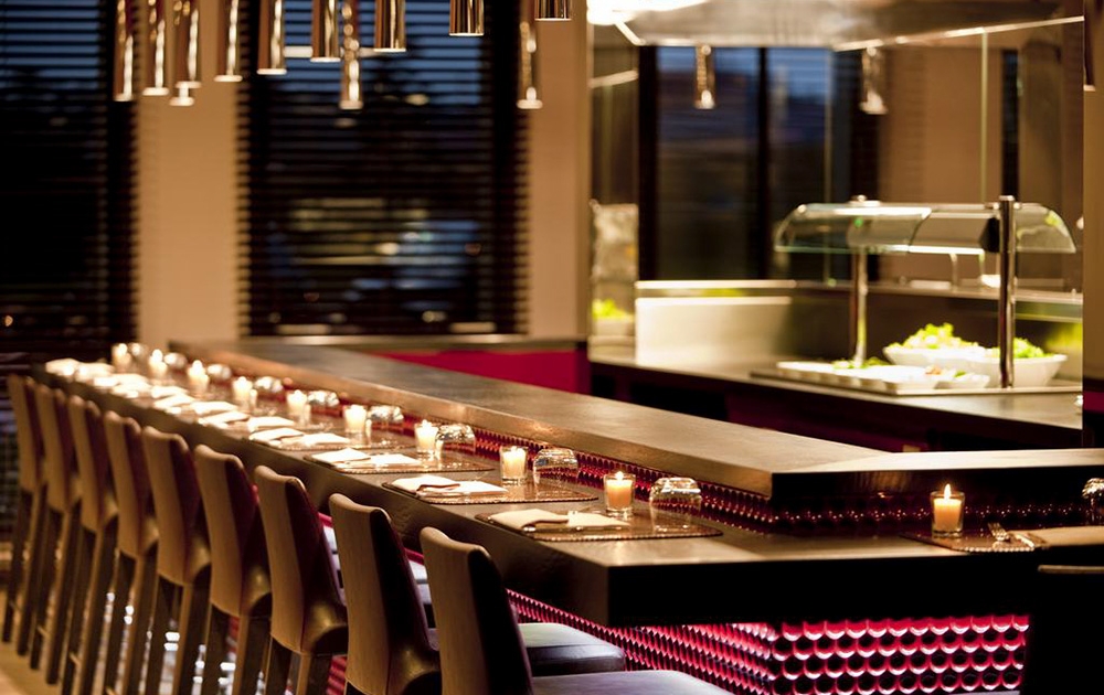 Un restaurant moderne et élégant avec un long comptoir noir épuré bordé de chaises à haut dossier et faiblement éclairé par des suspensions. Des bougies sont placées le long du comptoir et un bar à salades est visible en arrière-plan, ajoutant à l'ambiance chaleureuse.