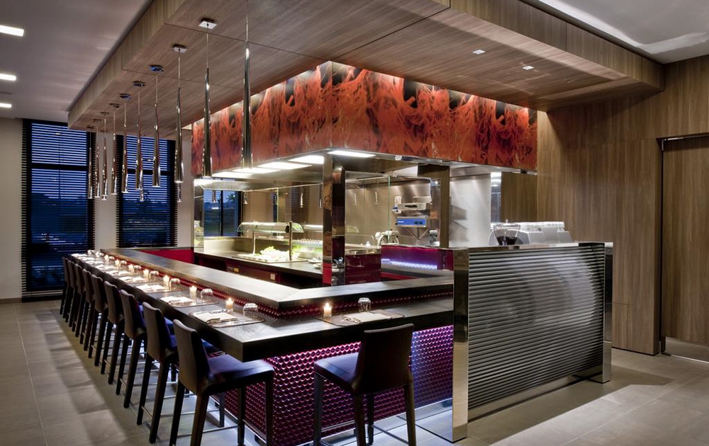 L'intérieur d'un restaurant moderne et haut de gamme comprend une cuisine centrale ouverte avec un design en acier inoxydable et en bois. Autour de la cuisine se trouvent de hauts comptoirs de salle à manger avec des chaises sombres soigneusement disposées, rappelant un bel agencement. Des suspensions sont suspendues au plafond et les grandes fenêtres laissent entrer la lumière naturelle.