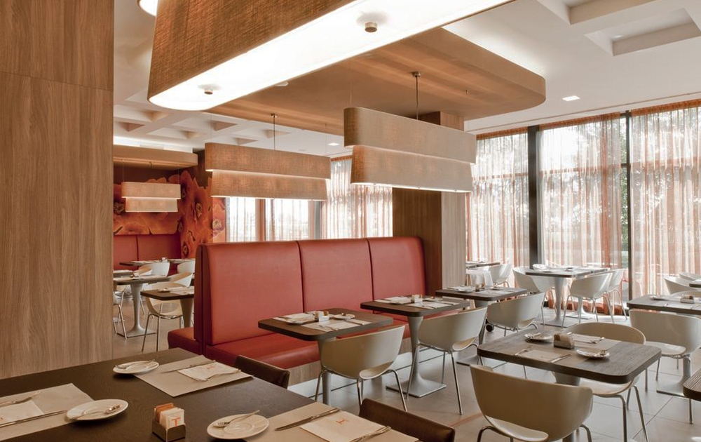 Un intérieur de restaurant moderne avec des banquettes tapissées de rouge et des chaises grises disposées autour de tables en bois foncé sur mesure. De grandes suspensions rectangulaires sont suspendues au plafond et des baies vitrées recouvertes de rideaux transparents permettent à la lumière naturelle de remplir cet espace luxueux.