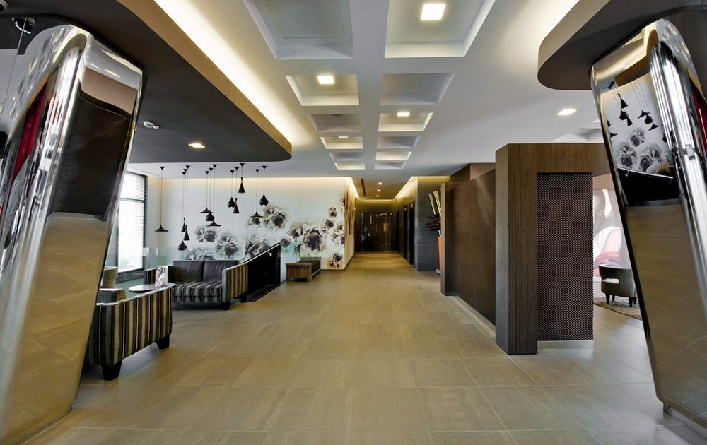 Le hall d'un hôtel moderne au design épuré et minimaliste présente un agencement expert. La décoration comprend des plafonniers abstraits, un long couloir, des contrastes de couleurs sombres et claires avec des motifs muraux artistiques et des colonnes en métal brillant. Des sièges confortables sont visibles sur le côté gauche.