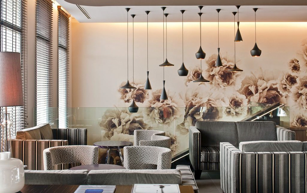Un espace salon moderne et élégant avec des chaises rembourrées grises aux motifs rayés et à motifs. Un groupe de suspensions noires est suspendu au plafond. Le mur du fond est décoré d'une grande fresque florale aux tons neutres, reflétant l'impeccable Aménagement hôtel. La lumière naturelle filtre à travers les stores.