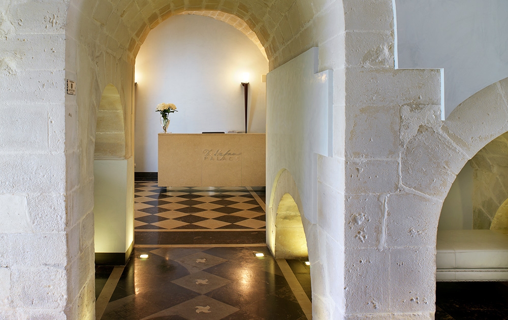 Un hall spacieux avec une arche en pierre menant à un comptoir de réception moderne, conçu avec soin grâce à un agencement bien pensé. L'espace est magnifiquement éclairé par un éclairage doux et le sol présente un motif en losanges noir et blanc saisissant. Des fleurs élégantes ornent le comptoir de réception en arrière-plan.