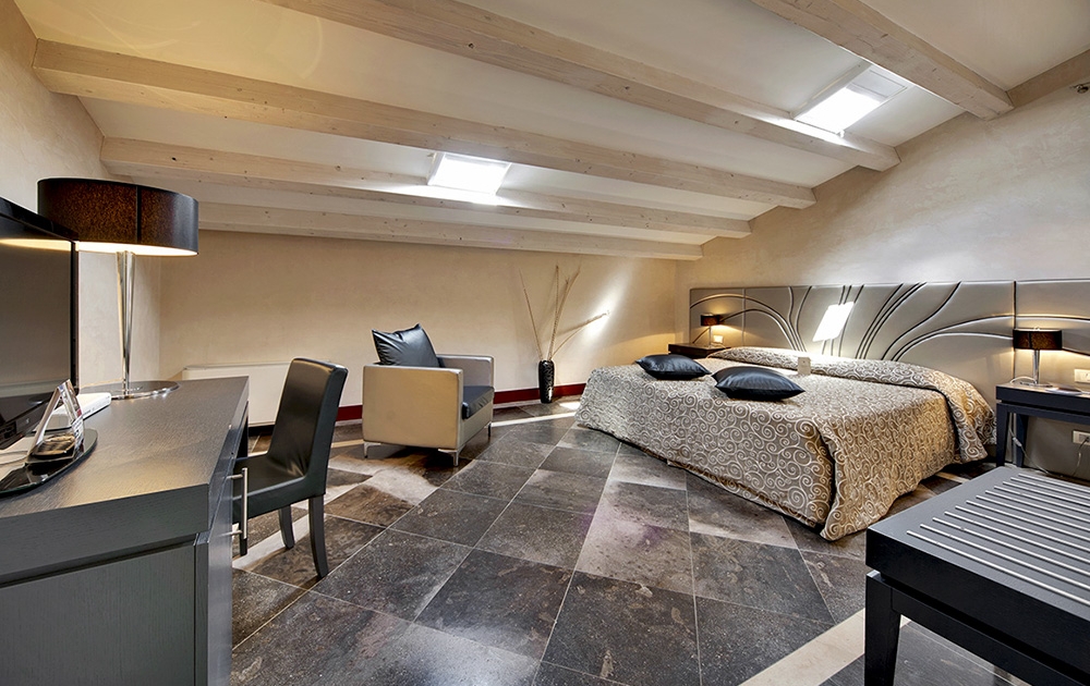 Une spacieuse chambre mansardée avec un plafond bas à poutres dégage une atmosphère luxueuse. La chambre comprend un lit double avec un couvre-lit à motifs, une chaise et une petite table, un bureau avec une lampe et une télévision à écran plat. Le sol est constitué de grands carreaux de pierre sombre. Des puits de lumière apportent de la lumière naturelle.