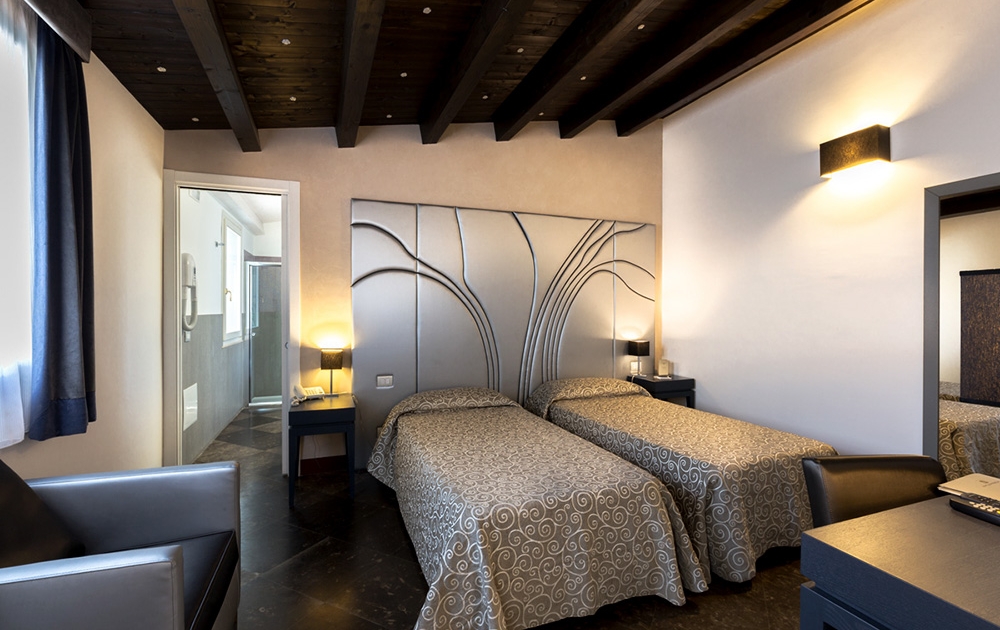Une chambre d'hôtel moderne dotée de deux lits simples avec couvre-lits à motifs, d'un coin salon avec une chaise noire, d'un bureau avec miroir et d'une porte menant à la salle de bain. Le design de l'hôtel Aménagement comprend des poutres en bois foncé et un éclairage d'ambiance pour une atmosphère chaleureuse.