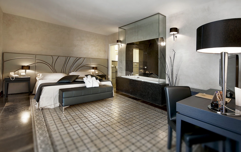 Une chambre d'hôtel moderne avec un grand lit, une tête de lit élégante et des tables de chevet avec lampes. Une salle de bain vitrée est visible avec une baignoire et un lavabo. Un bureau avec une chaise et une grande lampe noire est placé près de l'entrée. Le sol est doté d'un tapis à motifs, reflétant les options de conception de la boutique.