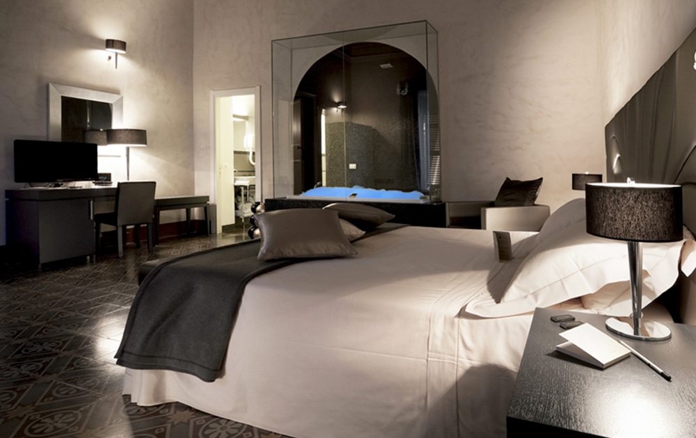 Une chambre d'hôtel moderne avec un grand lit recouvert de draps blancs et de coussins sombres. Le design élégant de la chambre comprend un éclairage tamisé, un bureau avec un ordinateur portable, une télévision à écran plat et une salle de bains vitrée. Des meubles sombres et une décoration élégante créent une atmosphère confortable.