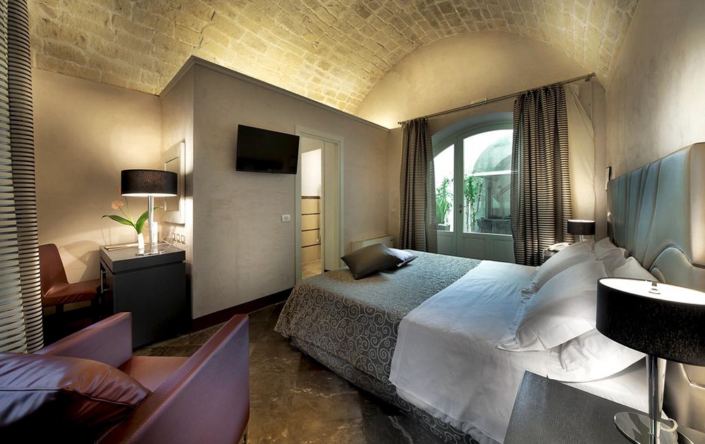 Une chambre confortable avec un plafond voûté en briques, comprenant un lit confortable avec une literie à motifs, une télévision à écran plat fixée au mur et un agencement bien pensé comprenant un bureau avec une lampe et une chaise. La fenêtre avec des rideaux rayés laisse entrer la lumière naturelle et il y a un petit fauteuil dans le coin.