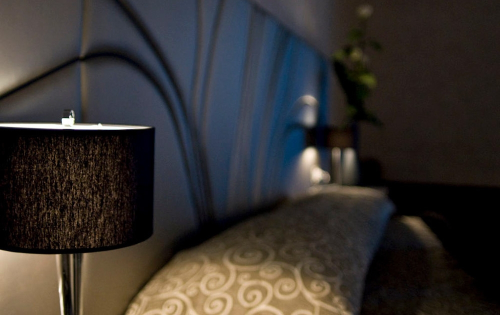 Une chambre à coucher moderne à la lumière tamisée présente un gros plan d'un abat-jour noir avec une lampe allumée projetant une douce lueur. La tête de lit présente un motif ondulé et le lit est recouvert d'une couette à motifs tourbillonnants. Une plante, faisant partie de l'agencement sur mesure, est partiellement visible à l'arrière-plan.