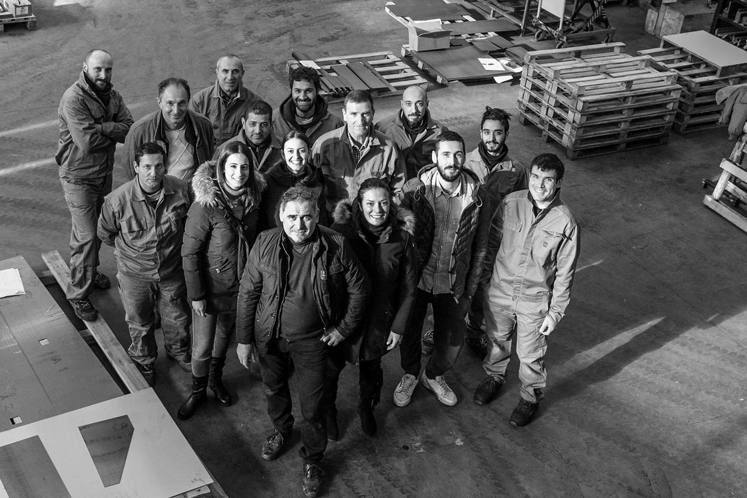 A black and white photo captures a group of 16 people standing in an industrial warehouse, smiling and looking up at the camera. The environment includes various tools, materials, stacks of wooden pallets, and elements from the contract division.