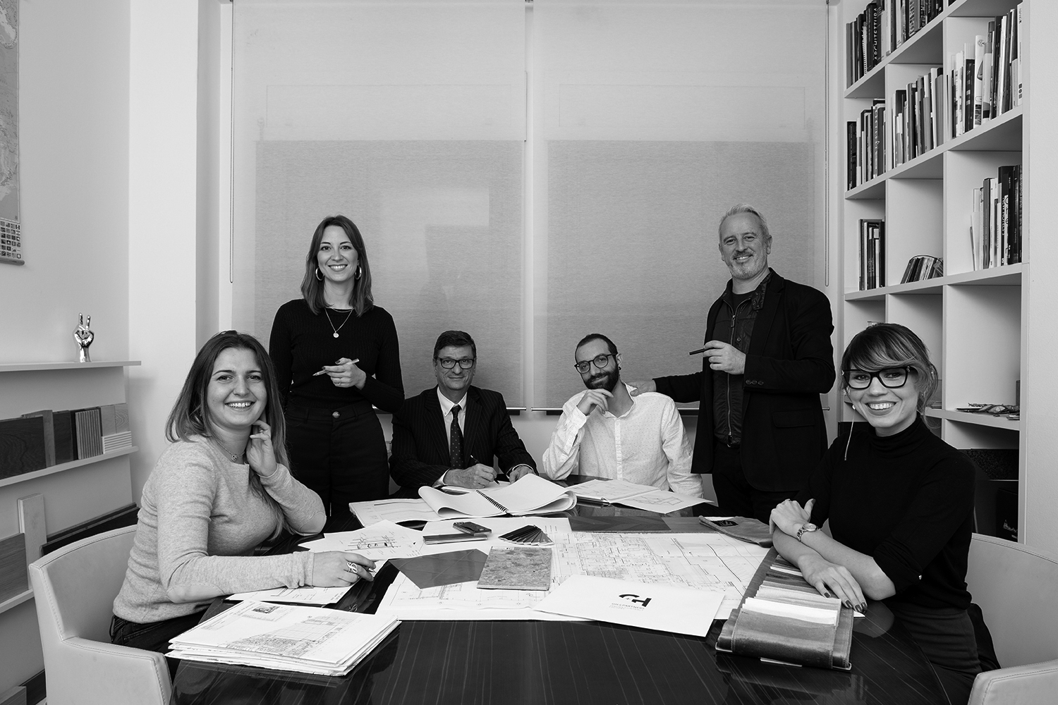 Black and white photo of six individuals at a table, engaged in a meeting or discussion. The table is covered with papers and documents. Bookshelves are visible in the background. Four people are seated, while two, reminiscent of a boutique hotel restaurant setting, stand behind them smiling and interacting.