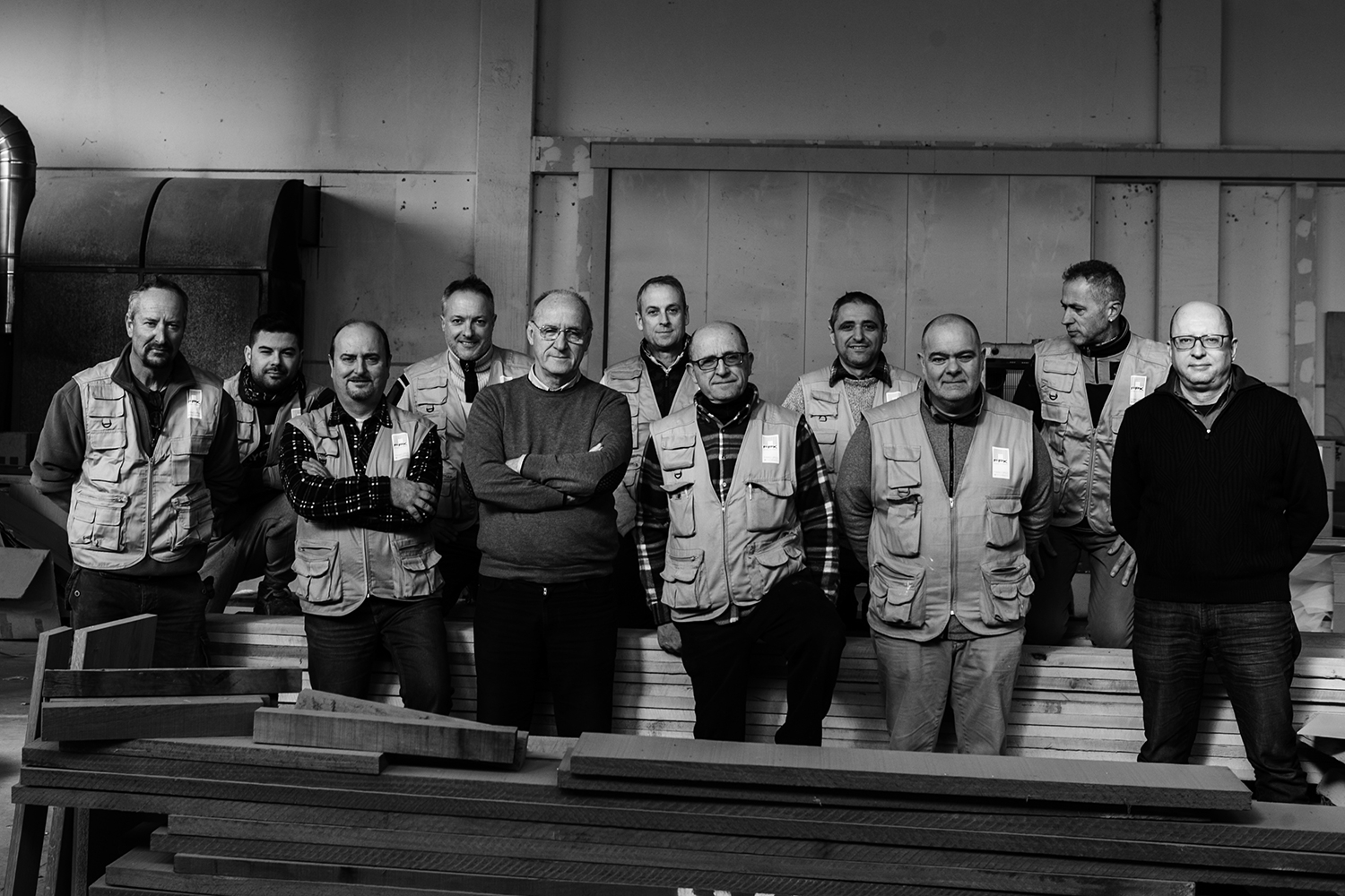 A group of eleven men, most wearing work vests, stand closely together in a workshop filled with wooden materials. Some wear glasses, and most have neutral or slight smiles. The workshop’s industrial interior is visible in the background, offering a boutique feel despite its raw setting. The image is in black and white.
