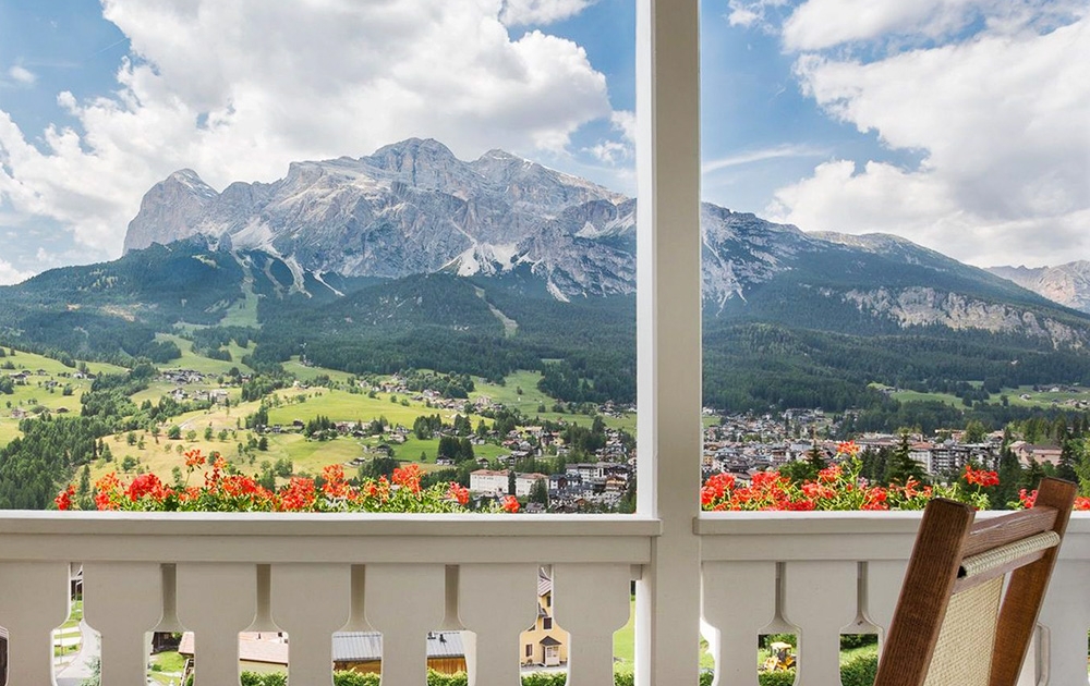 Une vue panoramique depuis un balcon présente une vaste chaîne de montagnes avec des sommets sous un ciel partiellement nuageux. En contrebas, une vallée verdoyante parsemée de maisons et d'arbres s'étend. Des fleurs rouges ornent la balustrade du balcon conçue sur mesure au premier plan, ajoutant une touche de couleur.