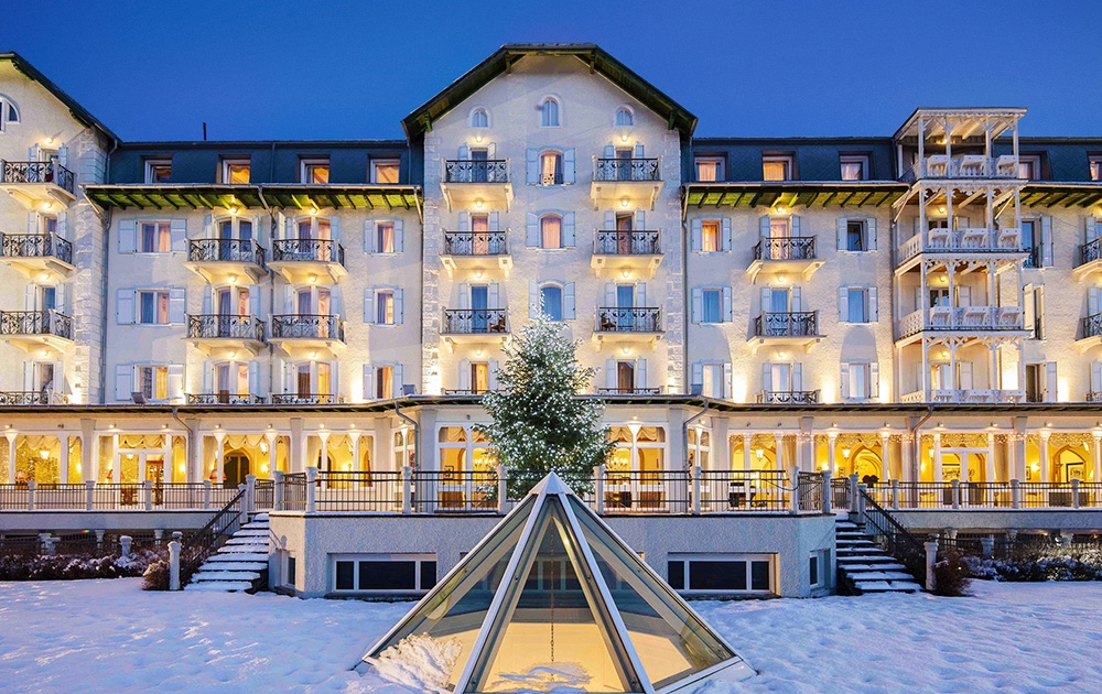 Un grand hôtel illuminé avec plusieurs balcons se dresse en arrière-plan sur un ciel crépusculaire. Au premier plan, un paysage enneigé entoure une petite structure en verre en forme de pyramide. L'aménagement luxueux de l'hôtel, associé à un éclairage jaune chaleureux, crée une atmosphère accueillante.