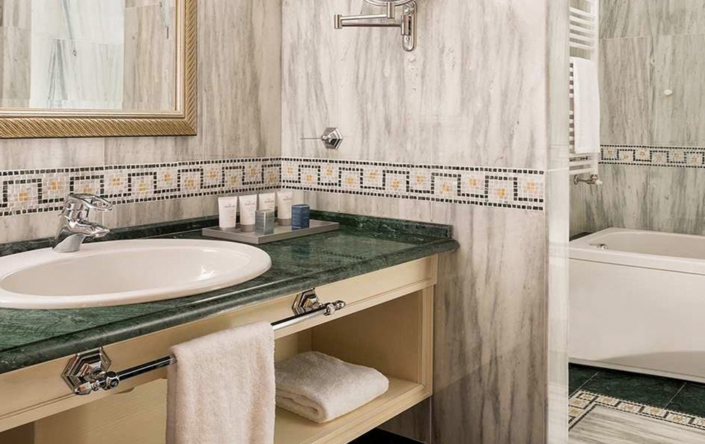 Une salle de bains moderne respire le luxe avec son plan de travail en marbre vert et son lavabo encastré. Au-dessus du lavabo, un miroir au cadre doré renforce l'opulence. Le mur est carrelé d'un motif en marbre gris, tandis qu'une serviette est élégamment suspendue à une barre chromée sous le comptoir. Parfait pour une division d'aménagement hôtelier contract, les produits de toilette sont soigneusement disposés sur le comptoir.
