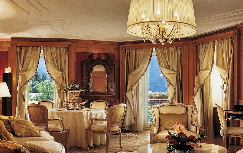 Un salon luxueux avec un élégant lustre suspendu aux hauts plafonds en bois. La pièce, au charme d'une boutique, dispose de grandes fenêtres drapées de rideaux de couleur crème. Il y a une table ronde recouverte de blanc, des fauteuils classiques et des décorations florales exquises pour cette touche sur mesure parfaite.