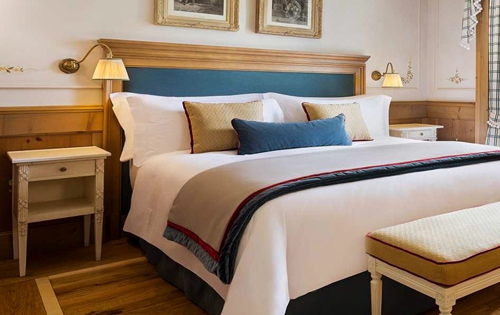 Une chambre confortable et de charme avec un lit soigneusement fait avec des draps blancs, des coussins bleus et beiges et une tête de lit rembourrée. Deux tables de chevet avec lampes encadrent le lit. Au-dessus de la tête de lit, deux tableaux encadrés ornent le mur. Un banc rembourré se trouve au pied de ce refuge sur mesure.