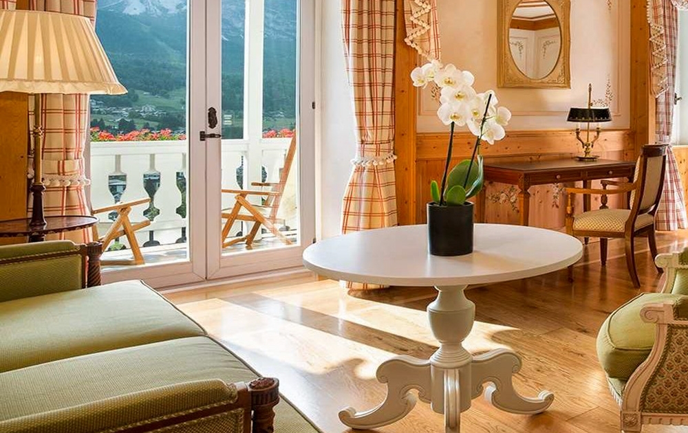Un salon luxueux avec une table blanche sur laquelle repose une orchidée en pot. Deux fauteuils et un canapé entourent la table. Un bureau avec miroir est contre le mur et des portes vitrées s'ouvrent sur un balcon avec vue sur la montagne. La pièce, conçue dans un style sur mesure, dispose de parquet et de rideaux à carreaux.
