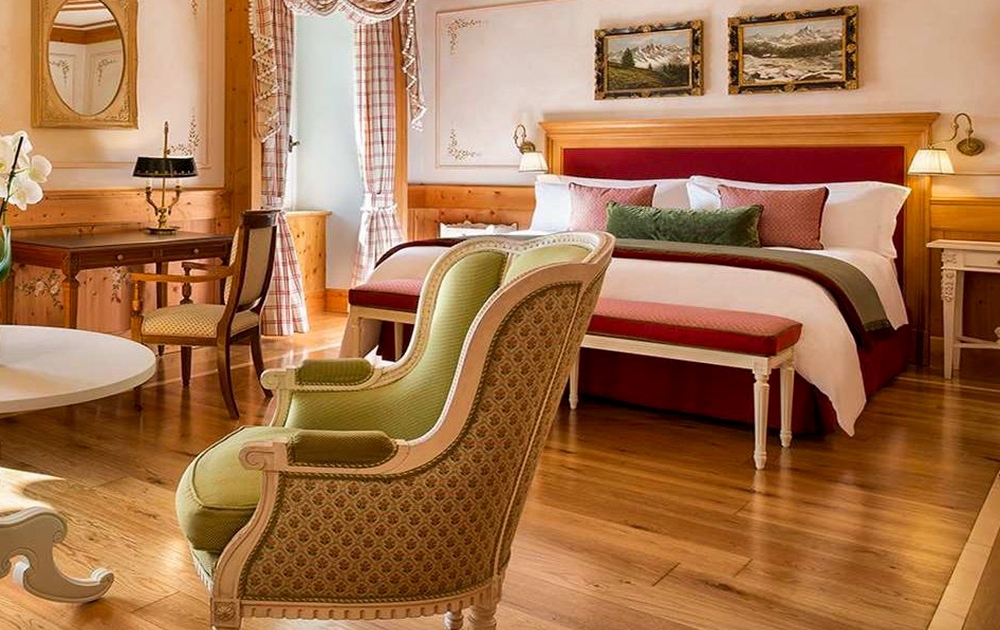 Une chambre d'hôtel confortable et élégante avec un grand lit orné d'oreillers et de couvertures. L'ameublement de l'hôtel comprend des boiseries, des meubles vintage, un bureau et deux fauteuils rembourrés. Des tableaux sont suspendus au-dessus du lit, tandis qu'une fenêtre avec des rideaux laisse entrer la lumière naturelle.