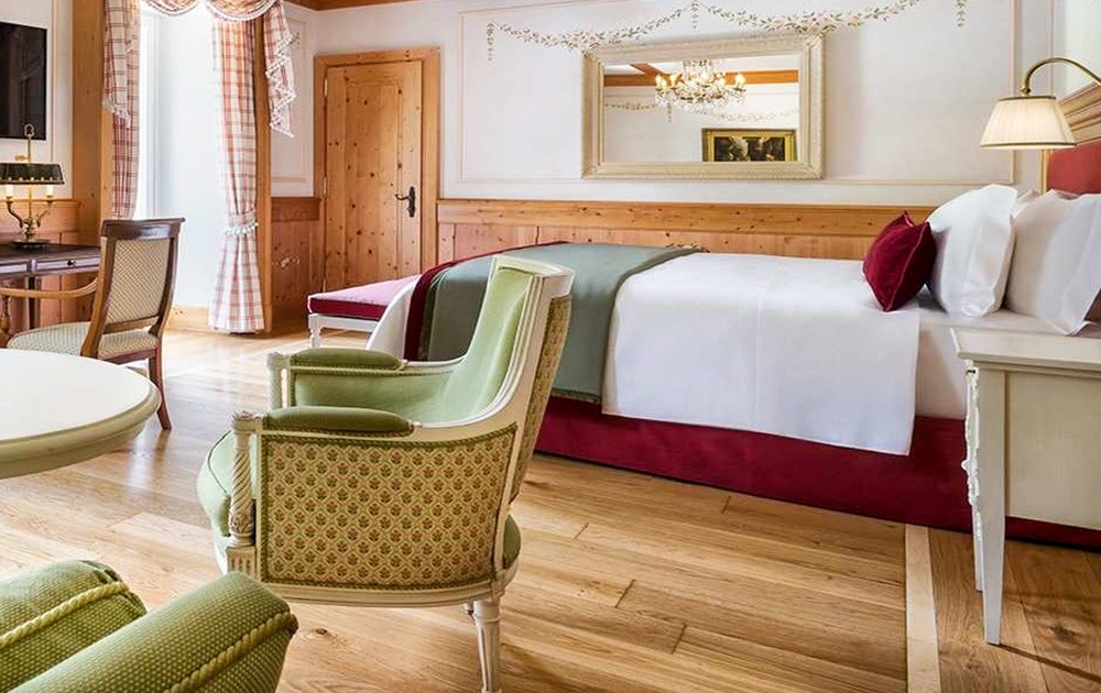 Une chambre d'hôtel spacieuse avec un lit soigneusement fait avec des draps blancs et des touches de rouge, un grand miroir mural au-dessus du lit, des fauteuils tapissés verts, un bureau avec une lampe et une fenêtre ornée de rideaux à carreaux. Le sol est en bois poli, mettant en valeur un aménagement hôtelier sur mesure impeccable.