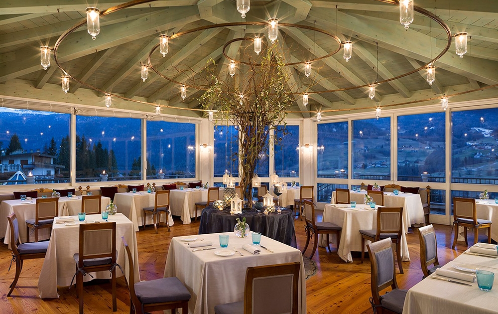 Un restaurant chaleureux avec un plafond en bois rustique décoré de lumières suspendues et d'un arbre en guise de centre de table, reflétant la touche sur mesure de notre division contract. La salle à manger comprend des tables habillées de verres et de chaises en bois, tandis que de grandes fenêtres offrent une vue panoramique sur le paysage vallonné à l'extérieur.