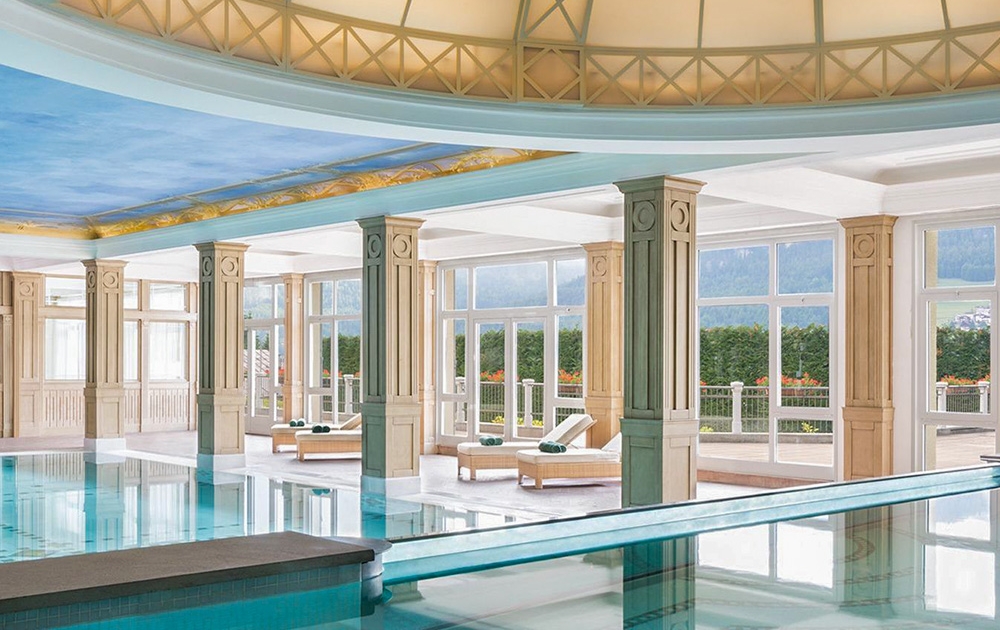 Espace piscine intérieur luxueux avec piliers élégants et grandes fenêtres offrant une vue panoramique sur la verdure et les montagnes. Le plafond, conçu par notre division de contrat de boutique, est orné d'un ciel bleu peint, et il y a plusieurs chaises longues avec des serviettes roulées pour se détendre.