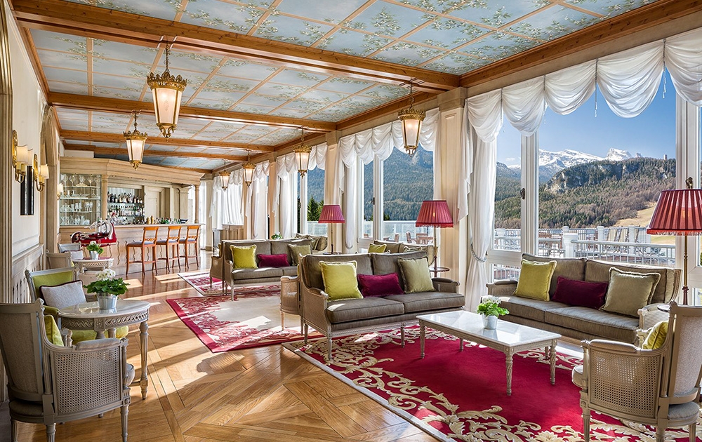 Salon élégant avec baies vitrées offrant une vue sur la montagne. La pièce comprend des sièges moelleux avec des coussins verts et violets, un tapis rouge et blanc orné, des lanternes suspendues et un plafond décoratif. Un espace bar avec tabourets est visible en arrière-plan, reflétant un agencement de luxe sur mesure.
