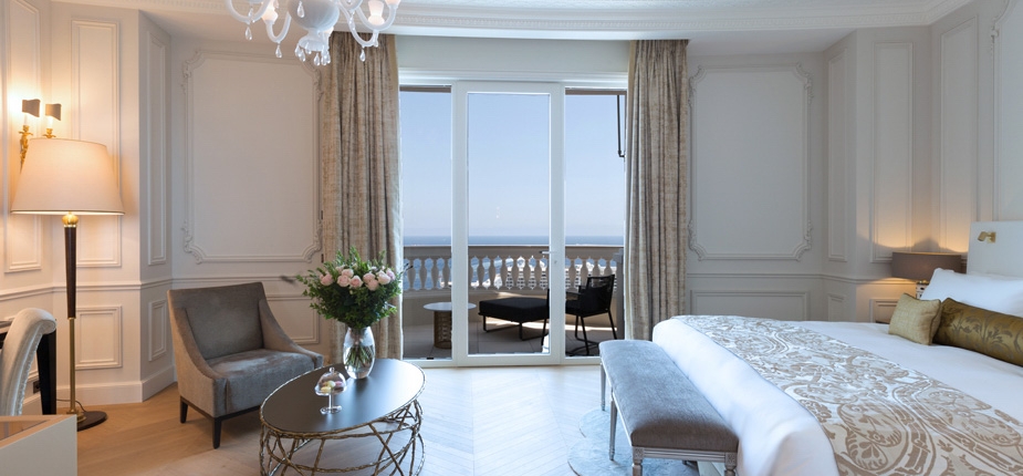 A luxurious bedroom with elegant decor features a king-sized bed, ornate bedding, and a pair of armchairs around a glass coffee table with flowers. Large French doors open to a balcony with a sea view. A chandelier hangs from the ceiling, enhancing the sophisticated boutique atmosphere of this luxe agencement.