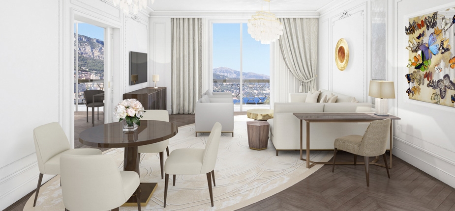 A spacious, elegantly decorated living room with a light color scheme. The room features a round dining table with four chairs, a cozy seating area, large windows with a mountain view, and modern art on the walls. A luxe chandelier hangs from the high ceiling, offering an amenagement hotel restaurant atmosphere.