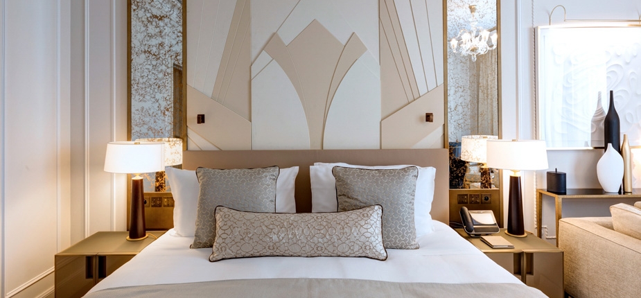 A stylish boutique hotel room features a neatly made bed with decorative pillows, framed by a modern white and gold accent wall. Nightstands on either side hold matching lamps. Other details include contemporary decor and wall-mounted lighting, creating a luxe ambiance.
