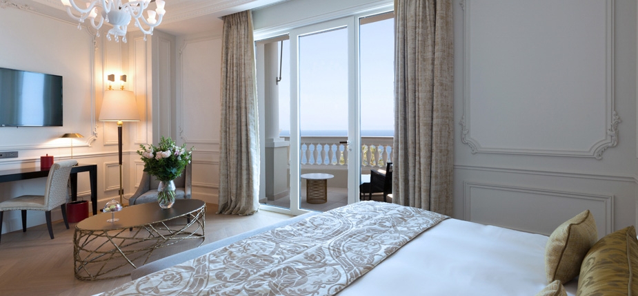 Luxurious boutique hotel room with elegant white decor, a large bed adorned with patterned bedding, a wall-mounted TV, a desk with a chair, and a contemporary coffee table with flowers. The open balcony offers an ocean view in this sur mesure retreat.