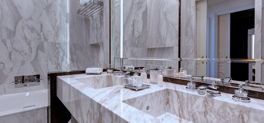 A luxurious bathroom with marble walls and countertops features a large, double sink vanity with chrome fixtures. The large mirror above the sinks is illuminated by elegant lighting. Sur mesure amenities, such as towels and toiletries, are neatly arranged on the countertop.