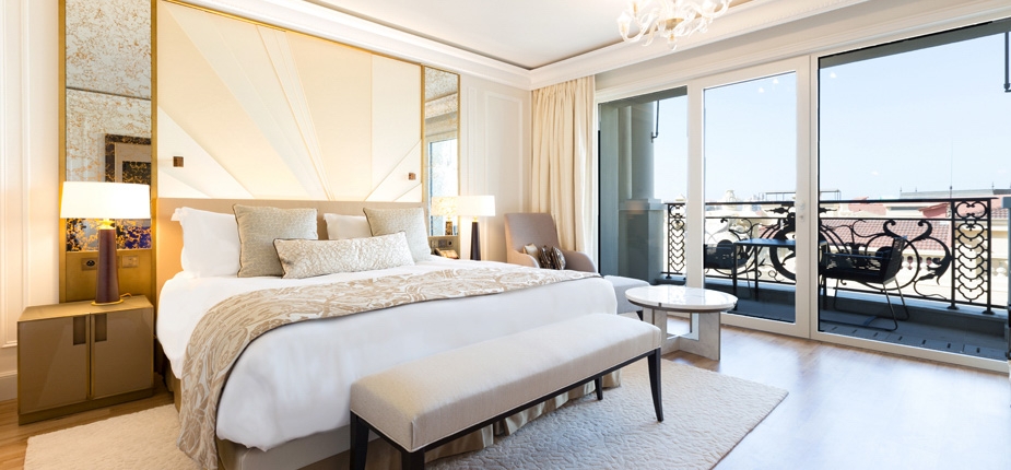 A luxurious, bright bedroom features a large bed with white linens and beige accents, a cushioned bench at its foot, and modern bedside tables with lamps. Designed by our contract division, the room opens to a balcony with outdoor furniture, accessible through sliding glass doors, and has a neutral color palette.