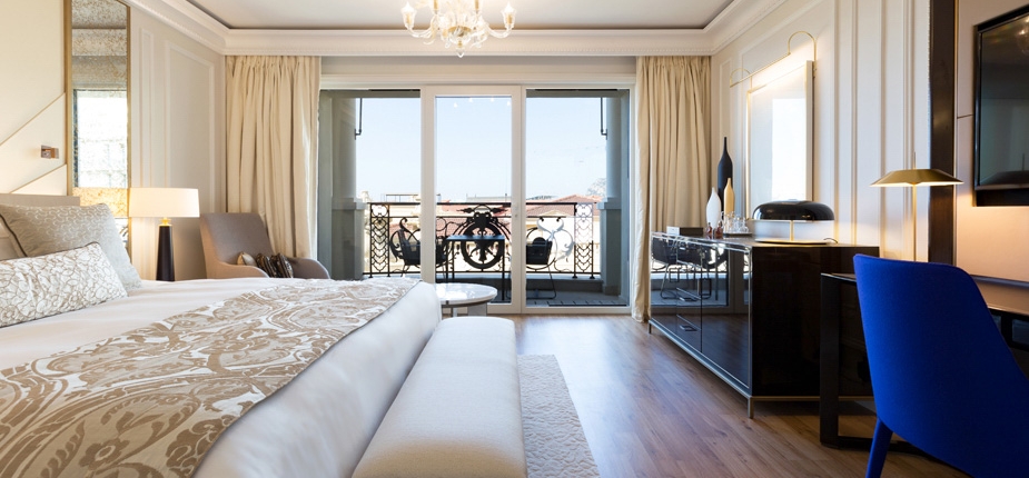 A luxurious hotel room with a large bed adorned with decorative pillows and a patterned bedspread. The room features a small desk with a blue chair, a flat-screen TV on the wall, and a chandelier. Floor-to-ceiling windows open to a balcony with scenic views, exemplifying true luxe living.