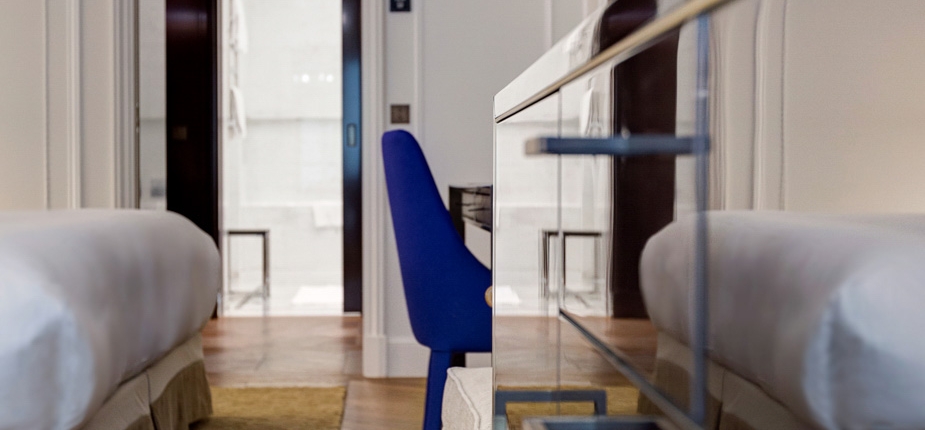 A modern hotel room with two beds on opposite sides of a central aisle. In the background, a bright bathroom is visible through an open door. A blue chair is placed at a desk between the beds, and a glass panel reflects part of the luxurious amenagement of the space.
