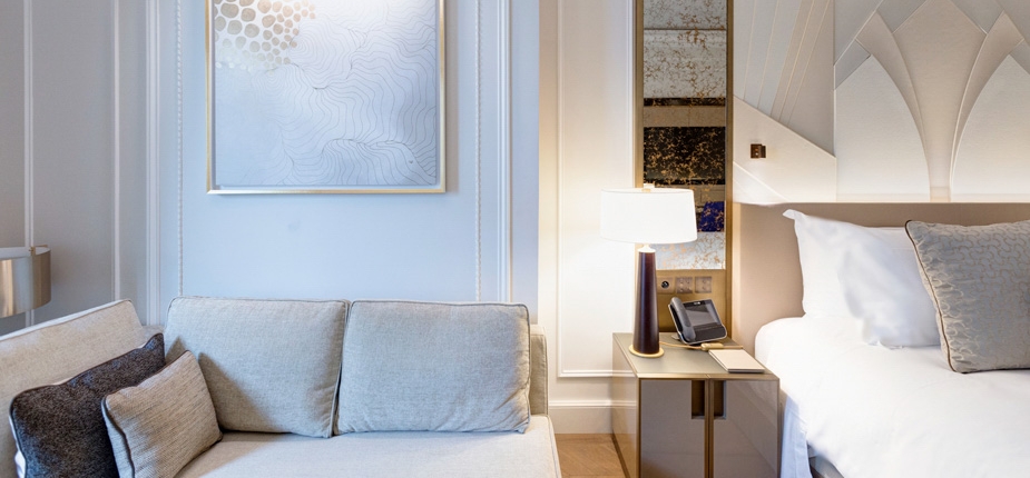 A cozy boutique hotel room features a soft couch with several pillows on the left, a bedside table with a lamp and a telephone in the center, and a bed with white linens and a decorative headboard on the right. A piece of abstract art hangs on the wall above the couch.